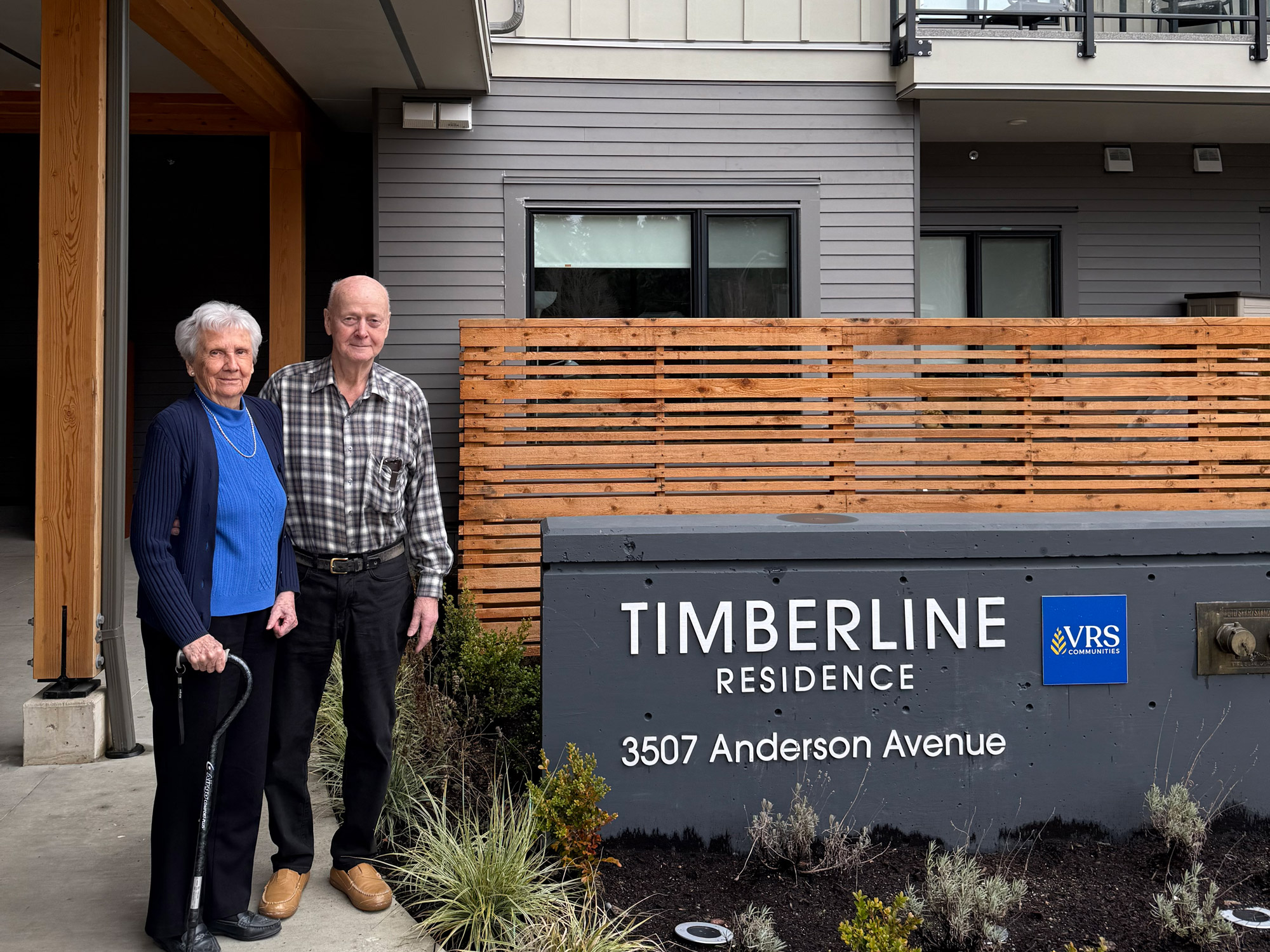 Bill and Edwina, two seniors standing in front of the Timberline seniors community sign